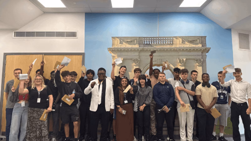 Learners from MK College receiving their T Level results. The students are stood in a line with two rows. They are smiling and holding up their results.