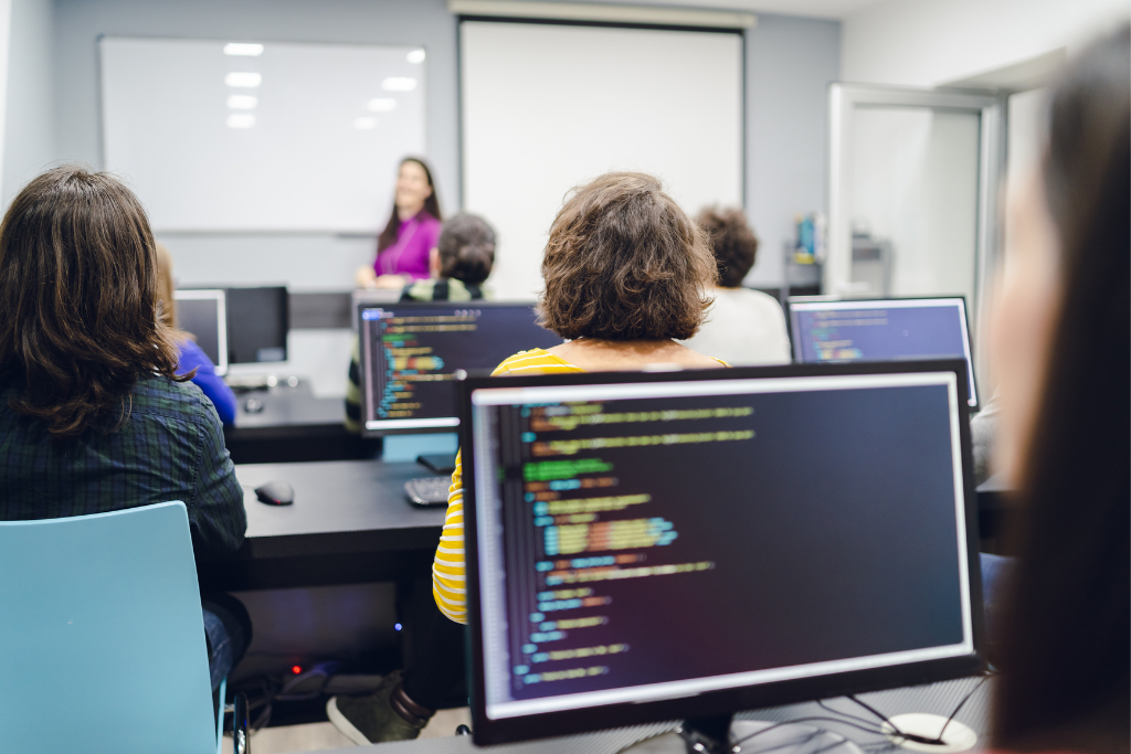 Classroom of students sat behind computers while a teacher stands at the front of the class teaching the lesson