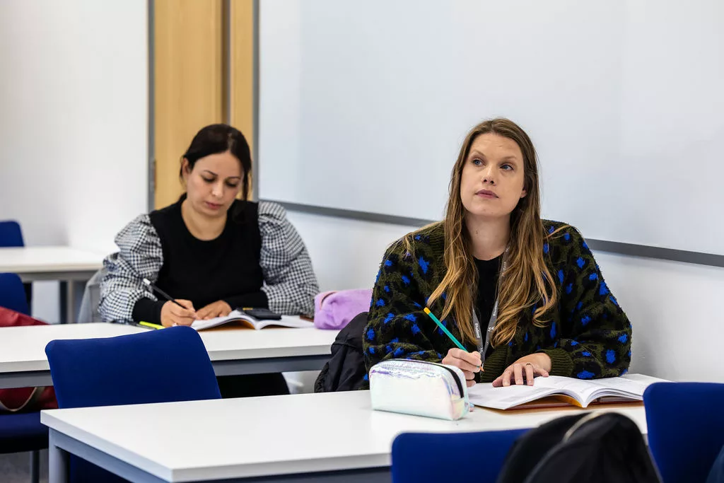 2 adults students sat behind desks working in notebooks