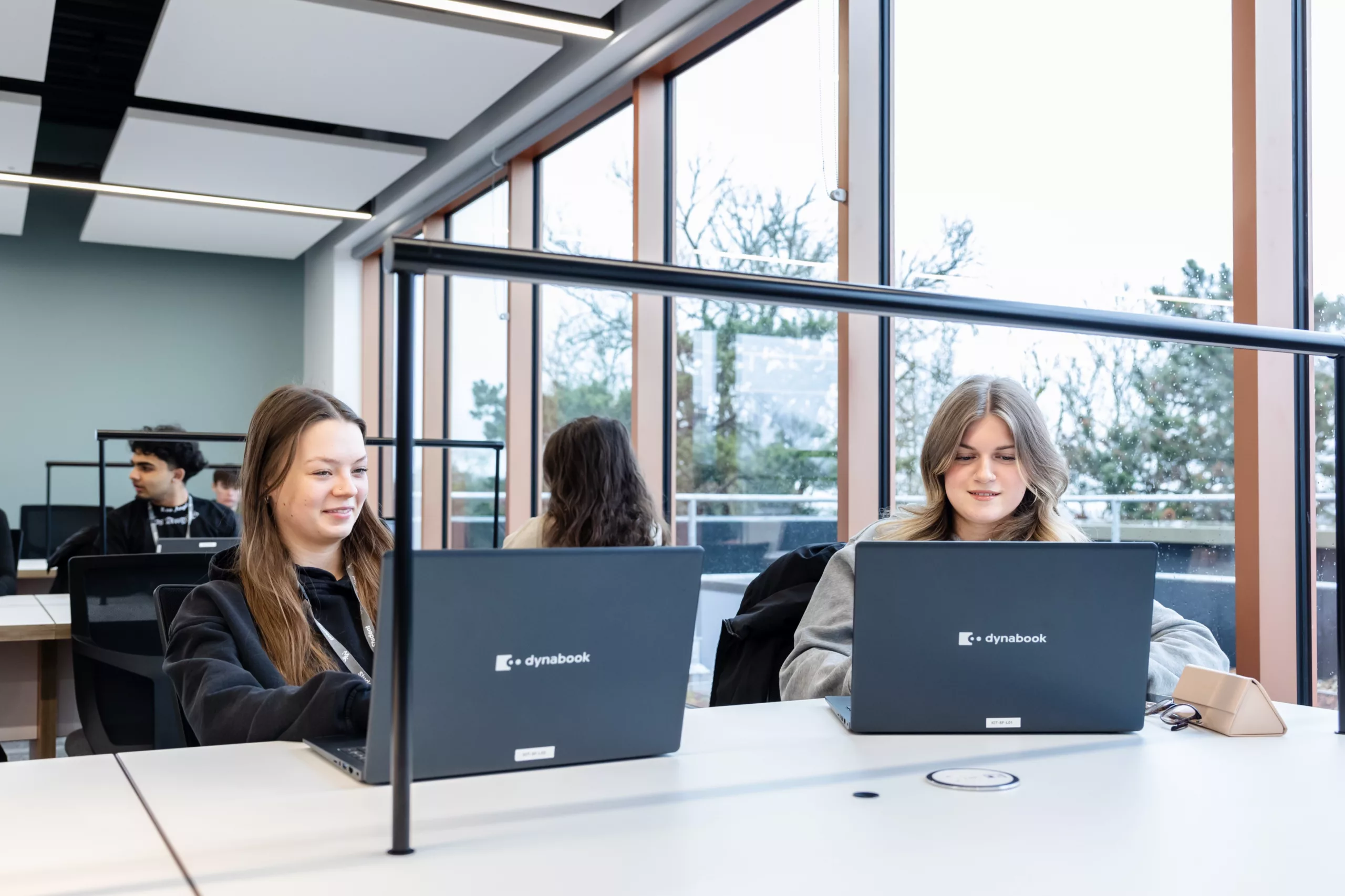 Students sitting at a modern study area with large windows, working on laptops.
