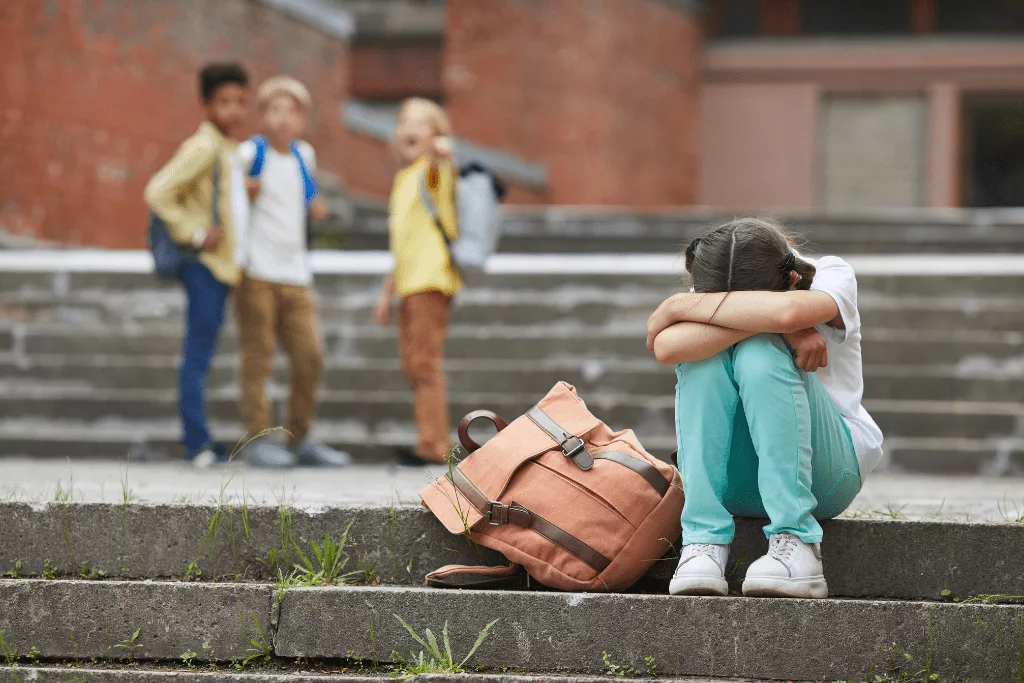 A student sitting next to her backpack on steps outside, crying or upset. A group of three children are standing in the background with one of them pointing at the upset student.