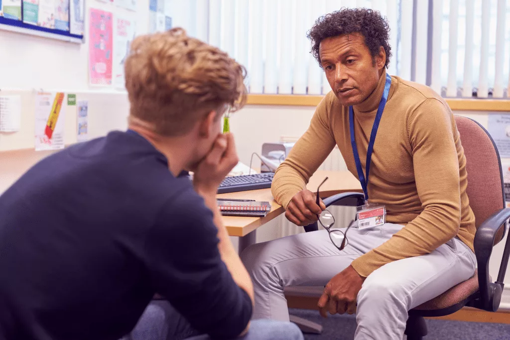 A teacher taking his glasses off while speaking to a student in an office.