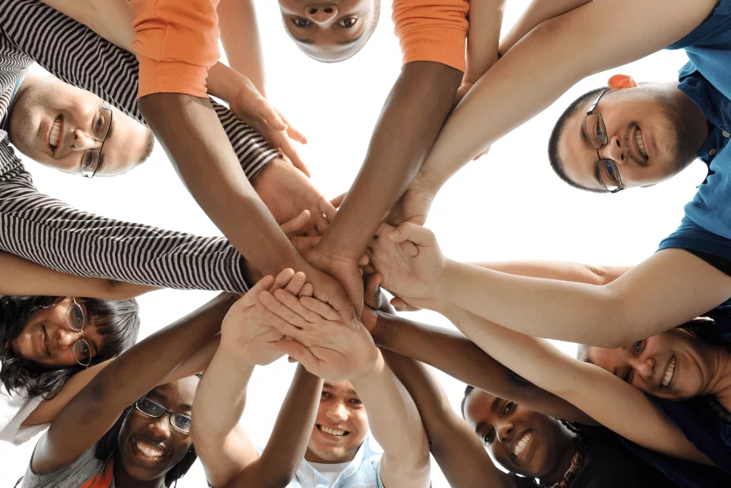 A photograph from below of people leaning forward and reaching their hands to the middle, smiling at the camera.
