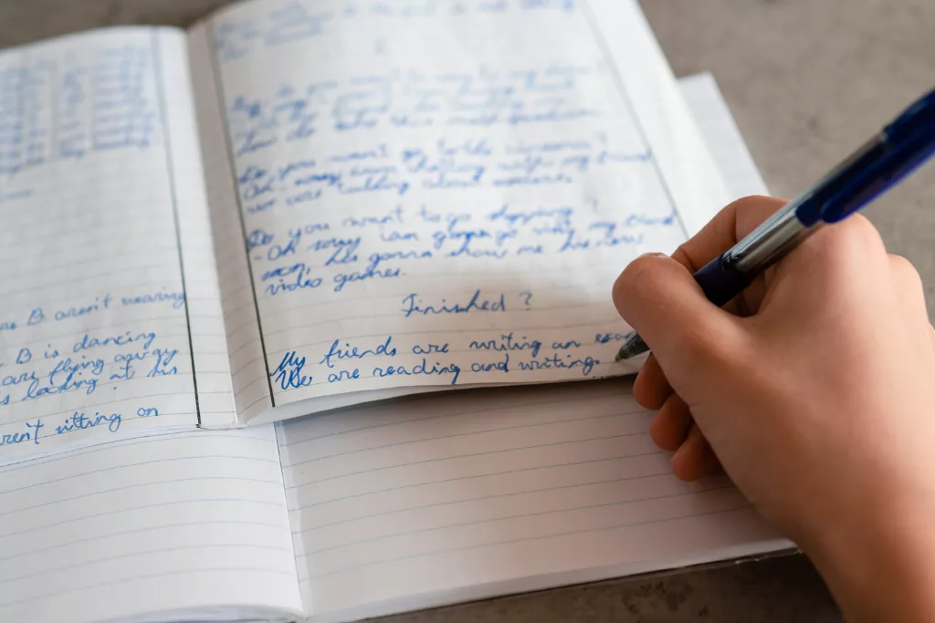 A photograph of a hand with a pen. The person is writing a story on a notebook with a blue pen.