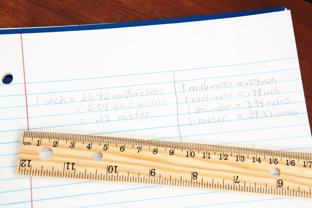 A photograph of conversions of inches to millimeters and vice versa on a lined notebook. A wooden ruler is laid across the page.