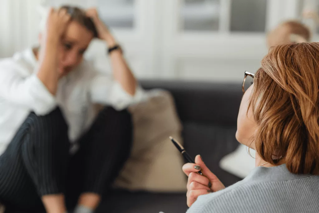A woman tilting her head and pointing a pen at a man in the background who is sitting down and holding his head, distressed.