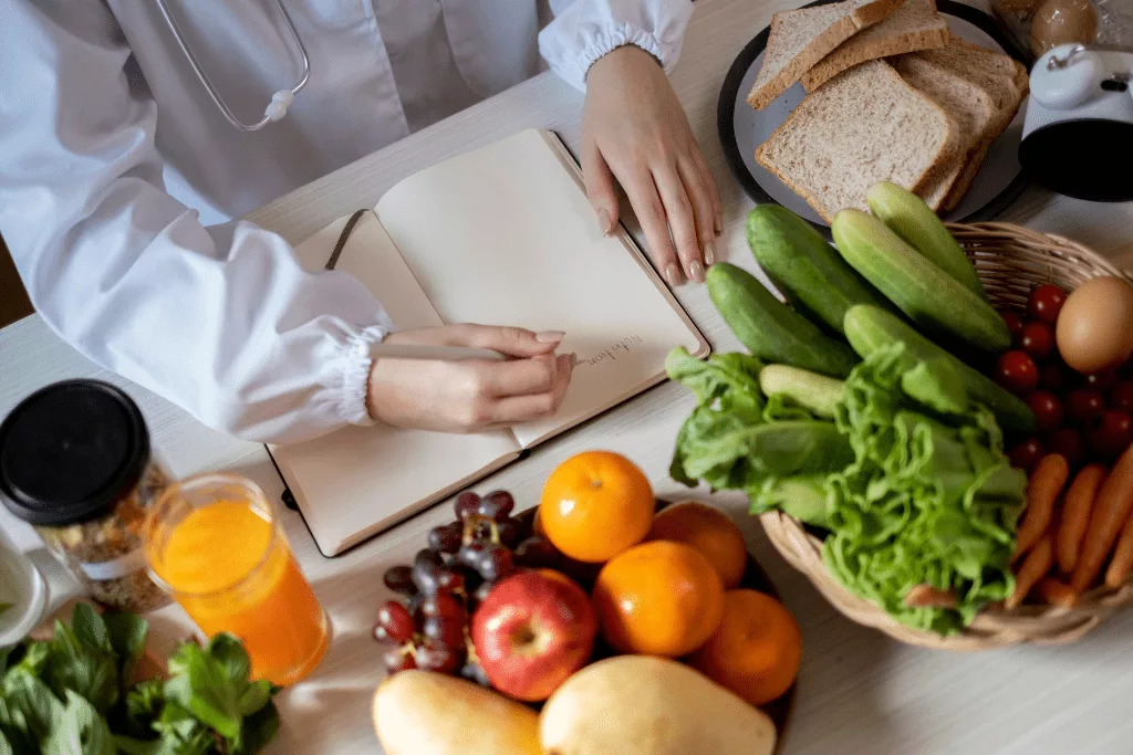 A photograph taken from above of a medical professional writing on an empty page in a notebook. The notebook is in the middle of different foods: juice, a bowl of fruit, a bowl of vegetables and a plate of bread.