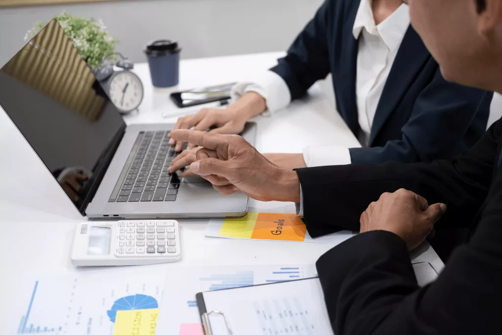 A photograph of two people wearing suits and sitting in front of a computer. On the table, there are papers, an alarm clock, a takeaway coffee cup and a calculator.