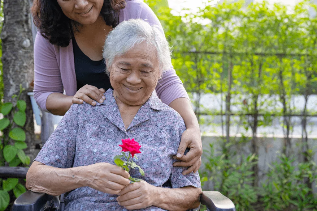 A younger woman leaning over an older woman who is holding a red flower and smiling.