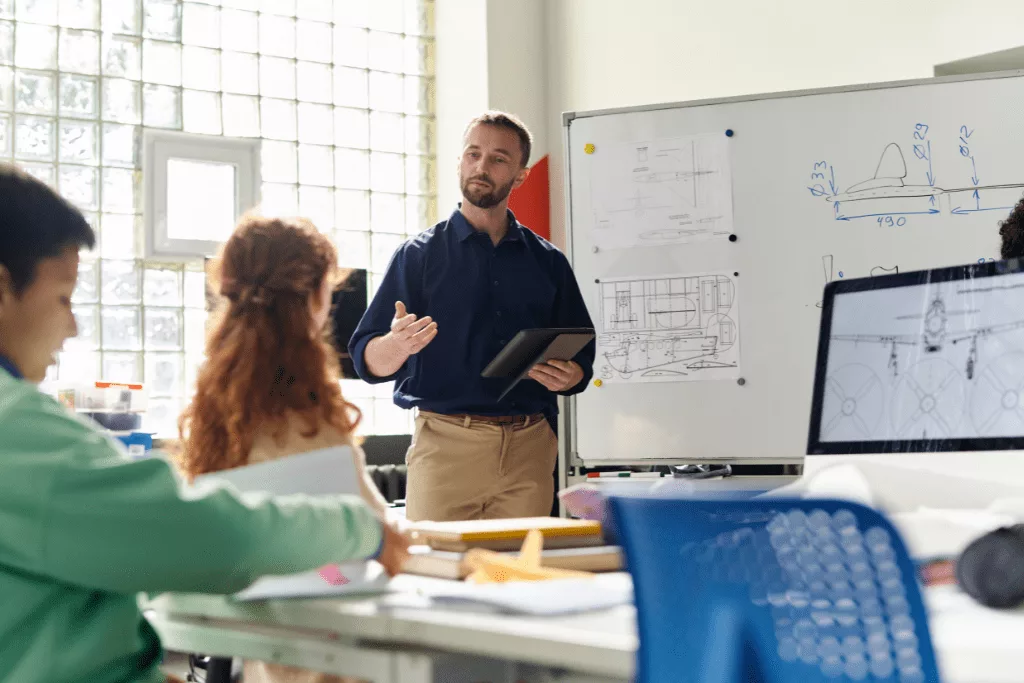A teacher speaking to two students, with graphs of an airplane on the whiteboard and computer screen.