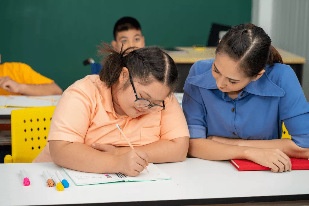 A woman sitting next to a student, looking at their work. The student is writing in their notebook.