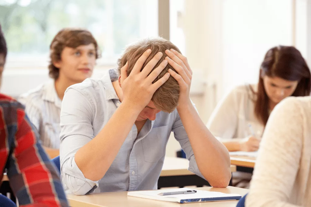 A confused and overwhelmed student holding his head with both hands, sitting at a desk and staring at his notebook.