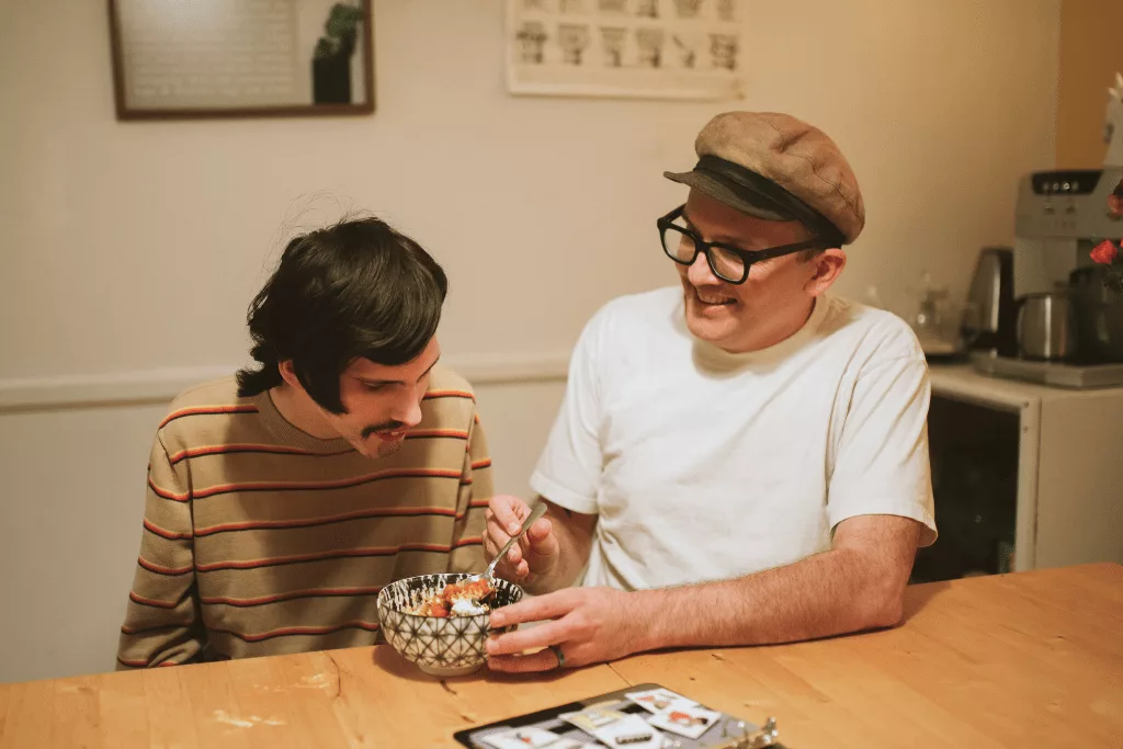 A man wearing a hat and glasses sitting next to a younger man, assisting him with eating.