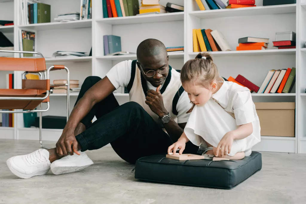 A man sitting on the floor in front of a bookshelf, next to a young girl who is sat on a pillow and looking at a wooden puzzle.