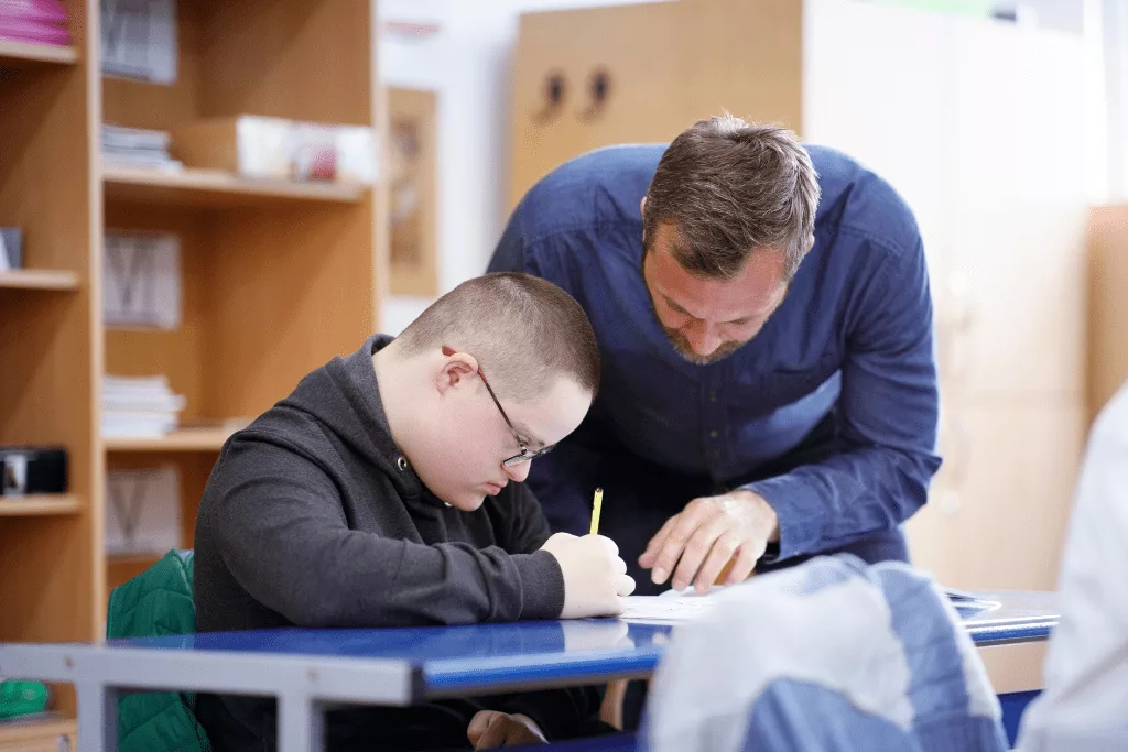 A man leaning over a desk, looking at the work of a student who is writing or drawing.