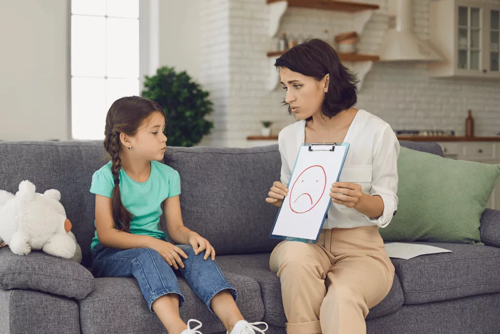 A woman with a empathetic expression showing a drawing of a sad face on a clipboard to a sad young girl.