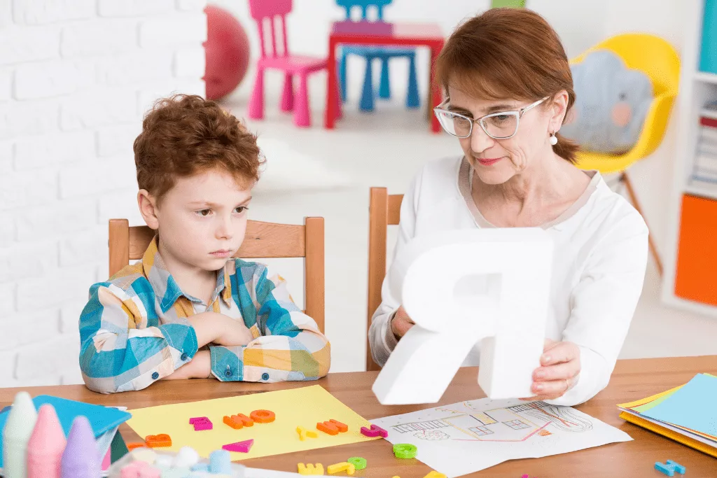 A woman holding a large letter R showing it to a young boy with his arms crossed. They are sat at a table with drawings, pens and plastic letters scattered over papers.