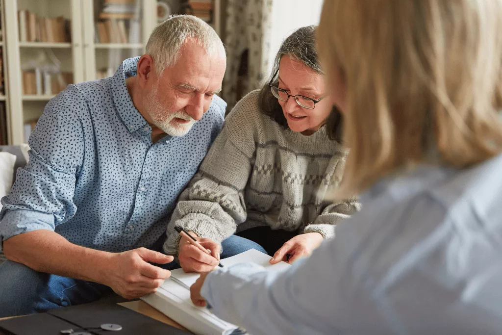 A younger woman pointing at a page in a book while an older couple look at the page, one of them holding a pen.